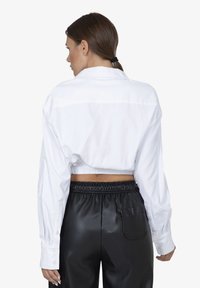 Camicia bianca a maniche lunghe, corta e con bottoni, con orlo elastico, abbinata a pantaloni neri in eco-pelle a vita alta con tasca laterale.