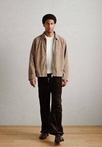 Brūns zamšādas žakete, krēmkrāsas krekls un tumšas kordurojas bikses. Modelis stāv uz koka grīdas; neitrāla fona dēļ apģērba detaļas izceļas.