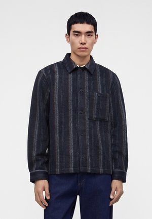 Ung mand iført en mørk, stribet knapjakke med en brystlomme foran og blå jeans, stående mod en simpel lys baggrund.
