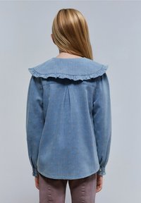 Ragazza con capelli biondi lisci che indossa una blusa blu a maniche lunghe con piccoli motivi e un ampio colletto arricciato, vista di spalle.