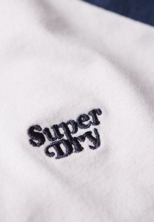 Tessuto bianco con primo piano di testo ricamato blu scuro che legge "Super Dry", mostrando una consistenza morbida e un design chiaro.