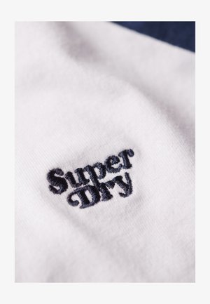 Biała tkanina zbliżeniowo przedstawiająca ciemnoniebieski haftowany tekst "Super Dry", ukazująca miękką strukturę i wyraźny wzór.