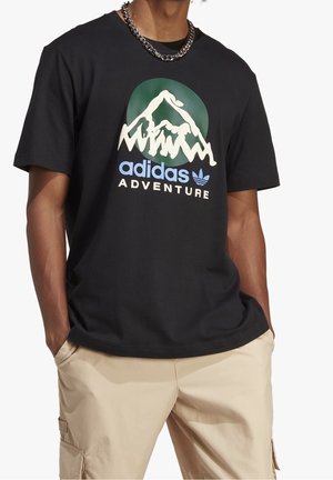 Schwarzes Adidas Adventure T-Shirt mit Berggrafik, getragen von einer Person mit beigen Cargo-Hosen und silberner Kette, Hände in den Taschen.