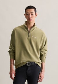 Jonge man in een olijfgroene sweatshirt met een kwart rits met het logo "MARC O'POLO DENIM" en zwarte jeans voor een effen achtergrond.
