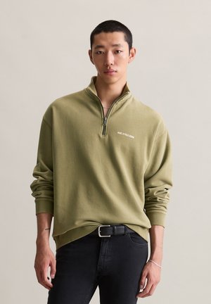 Jonge man in een olijfgroene sweatshirt met een kwart rits met het logo "MARC O'POLO DENIM" en zwarte jeans voor een effen achtergrond.