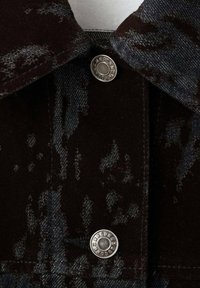 Giacca in denim nero con finitura texturizzata, bottoni in metallo argentato e colletto con un motivo sottile. Primo piano dei dettagli di chiusura.