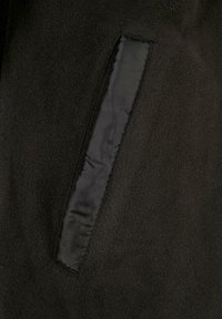 Nærbilde av svart fleece-stoff som viser en svart satinpocket trim. Teksturen er myk med en matt finish, som øker det visuelle interessen.
