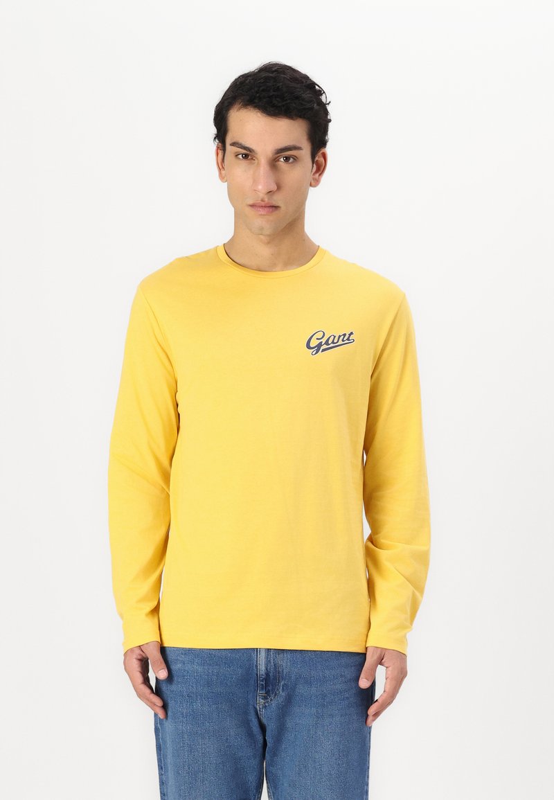 Langärmliges gelbes T-Shirt aus Baumwolle mit Rundhalsausschnitt und kleinem blauen Logo auf der linken Brust. Kombiniert mit blauen Jeans.