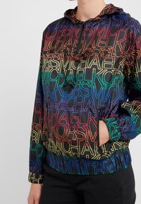 Giacca a vento con cappuccio a gradiente arcobaleno, caratterizzata da un motivo logo ripetuto in giallo, rosso, blu e verde su uno sfondo nero lucido.