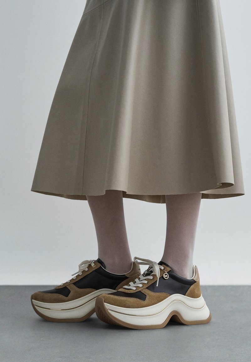 Vue en longueur des jambes d'une personne portant une jupe beige et des baskets marron, noires et blanches épaisses avec des semelles épaisses, debout sur un sol gris.