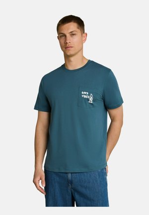 Mann trägt ein türkisfarbenes kurzärmeliges T-Shirt mit Vorderfach, auf dem weißer "Good Vibes"-Text und ein Surfbrettergrafik abgebildet sind, kombiniert mit blauen Jeans.