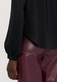 Blusa preta, texturizada, com mangas bufantes e detalhe de botão no punho, combinada com calças de pele sintética bordeaux com costura distinta nos bolsos.
