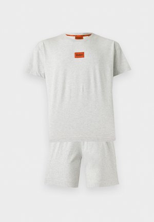 Mand stående barfodet iført lysegrå shorts og matchende T-shirt med lille rødt "HUGO"-logo på brystet, hænderne i lommerne.