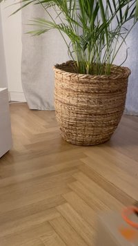 Naturalna pleciona kosz z zaokrąglonym kształtem w kolorze beżowym, zawierający zielone liście palmy, ustawiony na drewnianej podłodze w jodełkę.