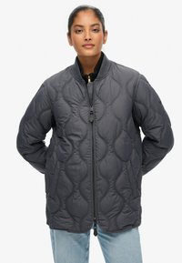 Giubbotto bomber trapuntato grigio scuro con zip frontale, collo a costine e tasche laterali. Presenta una texture liscia e sottili motivi ondulati.