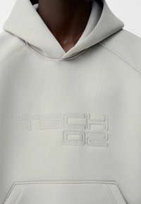 Ljust grå hoodie med en stor broderad "TECH 02"-logga. Har en dragsko i huvan och en mjuk, len textur med synliga sömmar.