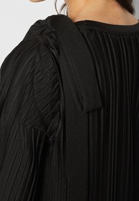 Musta laskostettu toppi, jossa on teksturoitu kangas. Ominaisuuksiin kuuluvat leveä, pehmeä olkaimen hihna ja herkät laskokset, jotka ulottuvat selän ja hihan varrelle.