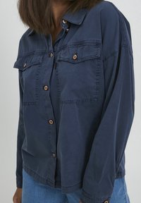 Navy blue button-up shirt with two chest pockets, metal buttons, and a relaxed fit. Cotton fabric appears smooth with a slight sheen.