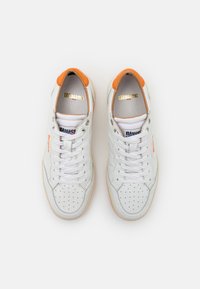 Des baskets en cuir blanc présentent des accents orange et des détails perforés, avec une semelle en caoutchouc beige et un design à lacets. Logo doré sur la languette.