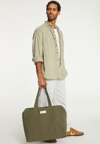 Sac en toile vert avec deux poignées, forme rectangulaire, et une étiquette blanche. Associé à un pantalon de survêtement gris clair et une chemise beige à boutons.