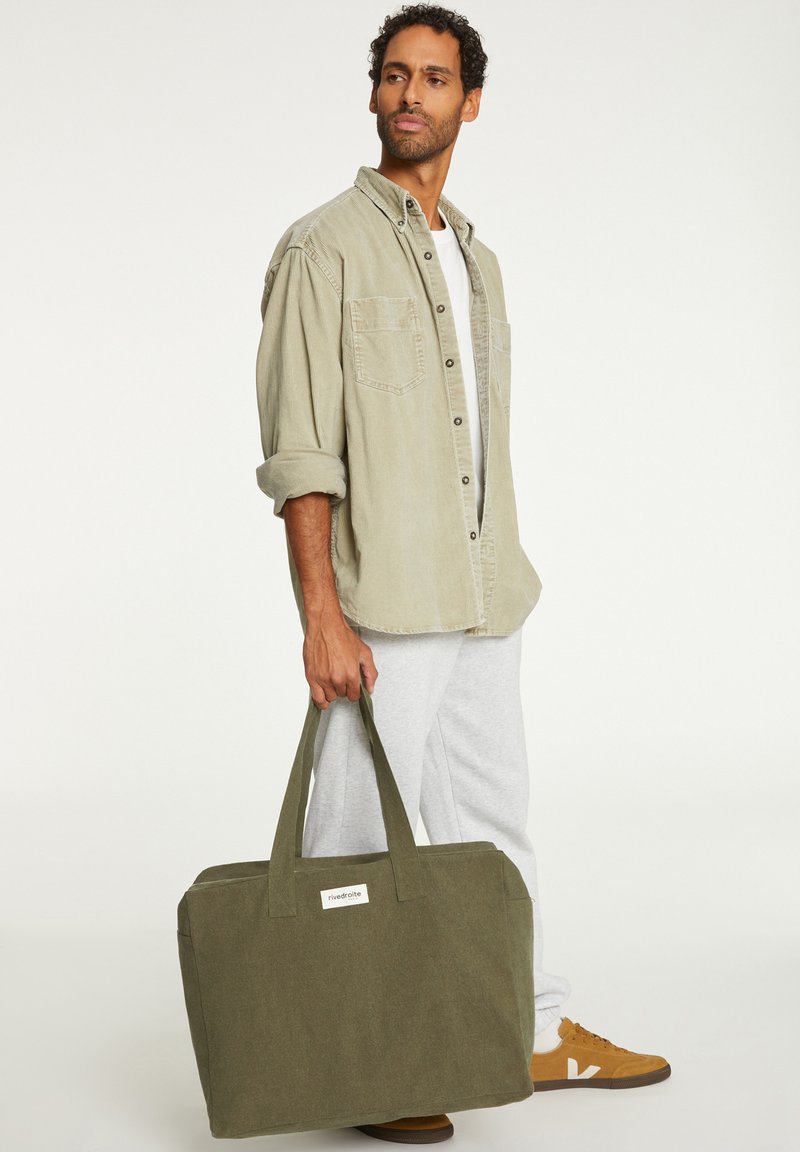 Sac en toile vert avec deux poignées, forme rectangulaire, et une étiquette blanche. Associé à un pantalon de survêtement gris clair et une chemise beige à boutons.