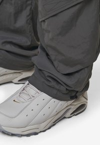 White leather sneakers with a textured surface and gold Nike logo; paired with dark gray, loose-fitting trousers featuring elastic cuffs.