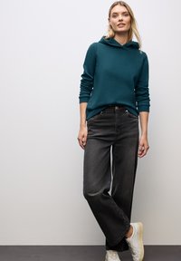 Felpa teal con tasca a canguro, abbinata a jeans neri a vestibilità ampia e scarpe da ginnastica bianche. L'outfit presenta un design casual e rilassato.