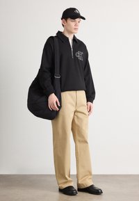 Černý pulovr na čtvrtový zip s logem, béžové široké kalhoty, černá taška a odpovídající kšiltovka, doplněné černými slip-on botami. Minimalistický design.