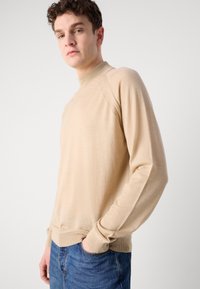Beige langermet genser med høy hals, ribbestrikkede ermer og kant, samt en glatt tekstur, kombinert med blå denimjeans.