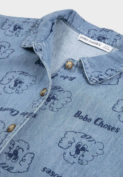 Chemise en denim bleu clair pour enfants avec fermeture à boutons, motifs de visages de chiens de dessin animé partout, et texte "Bobo Choses".