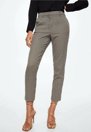 Femme portant un haut noir, un pantalon slim pied-de-poule à la cheville et des sandales à talons hauts à brides couleur camel, debout avec une main sur la hanche.