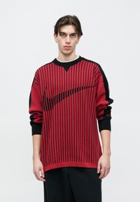 Piros és fekete kötött pulóver függőleges csíkokkal és nagy fekete logóval. Kerek nyakkivágás, hosszú ujjú, bordázott mandzsettákkal.