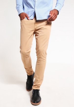 Mann trägt beigefarbene Slim-Fit-Hose, ein hellblaues Hemd mit Knöpfen und schwarze Schnürschuhe, geht mit den Händen in den Taschen vor schlichtem Hintergrund.