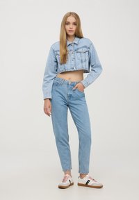 Giacca di jeans cropped azzurro chiaro con tasche frontali, abbinata a jeans azzurro chiaro a vita alta e sneaker bianche con accenti rosa e neri.