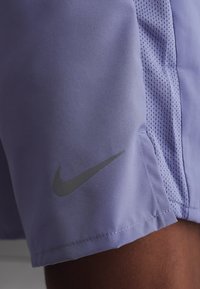 Ανοιχτό μοβ αθλητικό σορτσάκι με δίχτυ στο πλαϊνό μέρος και σκούρο γκρι λογότυπο Nike Swoosh κοντά στο τελείωμα.