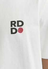 Hvid t-shirt med korte ærmer, der har sort tekst "RD" og "D" samt en rød prikkede. Lavet af blødt bomuld med en glat tekstur.