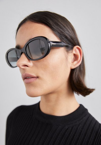 Black oval sunglasses with dark lenses, glossy frame, and subtle logo detail on temples. Worn by a person with short hair, against a neutral background.