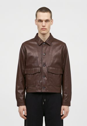 Un homme porte une veste en cuir marron foncé boutonnée sur le devant, avec deux poches à rabat, assortie à un pantalon noir sur un fond clair et uni.
