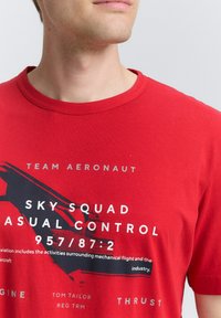 Camiseta de algodón roja con cuello redondo, texto impreso en negrita y diseño gráfico en blanco y negro en la parte frontal.