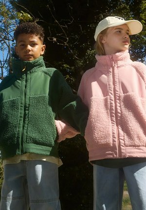Due bambini si tengono per mano, indossando giacche in pile: una verde con una finitura texturizzata e una rosa, entrambe dotate di colli alti e cerniere.