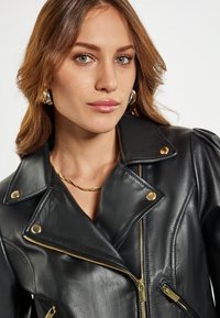 Giacca di pelle nera con hardware dorato, caratterizzata da un colletto a ribalta, chiusura frontale con zip e polsini con zip, e un design aderente. Texture liscia.