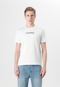 Baltas medvilninis marškinėliai su trumpomis rankovėmis ir apvaliu iškirpimu. Ant krūtinės yra violetinis „Calvin Klein“ logotipas.