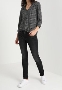 Frau trägt eine schwarz-weiß gemusterte Bluse, schwarze Skinny-Jeans und weiße Sneaker und steht vor einem schlichten, hellen Hintergrund.