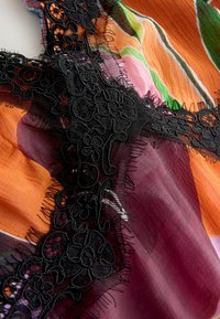 Tissu aux couleurs orange, vert, rose et bordeaux, orné d'une bordure en dentelle florale noire avec un tulle fin et des accents de franges.