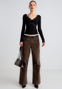 Μαύρο τοπ με μακριά μανίκια και δαντελένιες λεπτομέρειες, συνδυασμένο με φαρδιά, καφέ παντελόνια με ψηλή μέση. Το μοντέλο κρατάει μικρή καφέ τσάντα χειρός και φοράει μυτερά τακούνια.