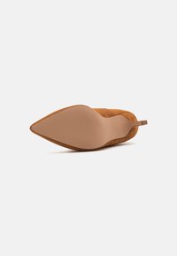 Tacco a punta in suede marrone con suola beige liscia e tacco piccolo; la parte posteriore include un anello per un facile indossamento.