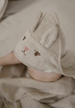 Copil înfășurat într-un prosop bej cu glugă, cu fața și urechile unui animal brodate, întins pe o suprafață moale.