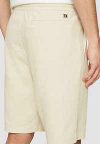 Pantalones cortos beige con una tela texturizada, que cuentan con una cinturilla elástica, bolsillos laterales y un bolsillo trasero adornado con un pequeño logo.