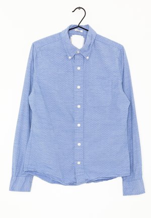 Hellblaues langärmliges Hemd mit kleinen weißen Punkten, einer Brusttasche und einem knöpfbaren Kragen auf einem schwarzen Bügel vor weißem Hintergrund.