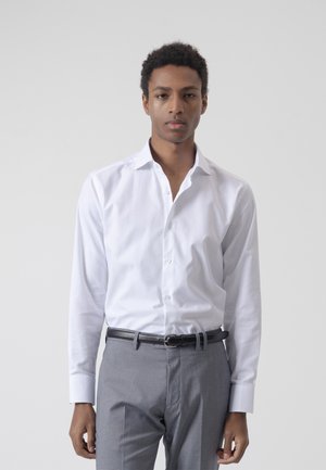 Joven con cabello corto y rizado, que lleva una camisa blanca abotonada y pantalones grises con cinturón negro, de pie frente a un fondo liso.